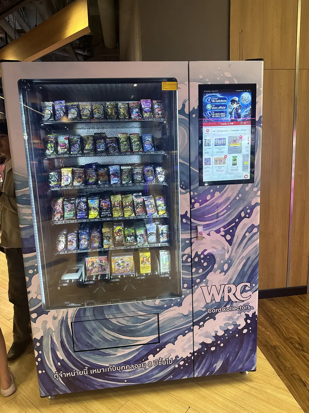 Framsidan av en TCG-automat i Bangkok med pekskärm högst upp och en belyst glasfront med numrerade fack fyllda med boosterpaket