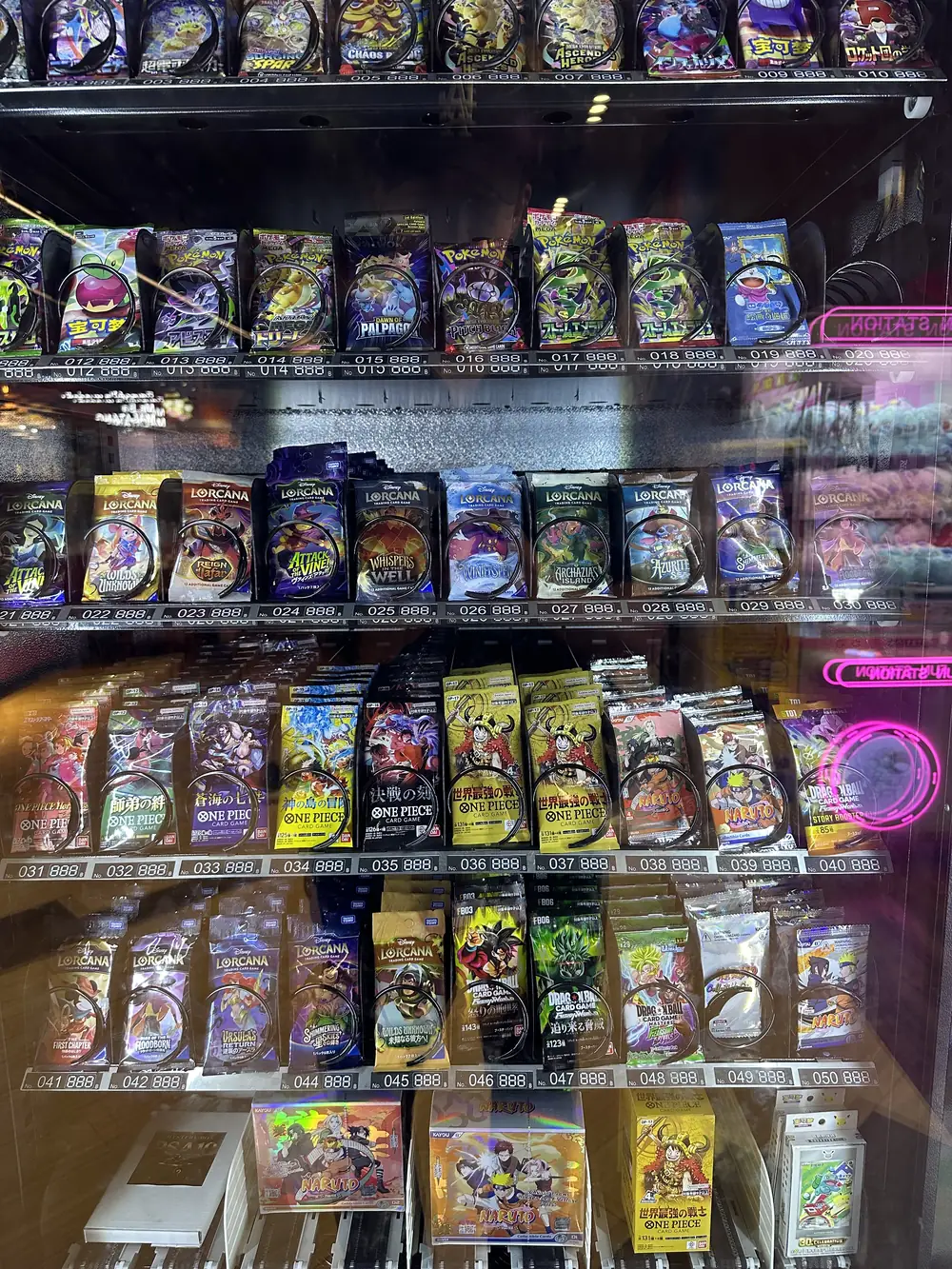 Förseglade One Piece-boosterpaket tillsammans med Lorcana, Dragon Ball, Naruto och Pokémon i numrerade fack bakom glasfronten på en TCG-automat i Bangkok